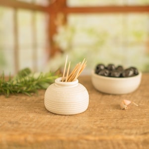 May include: White ceramic toothpick holder with a ribbed design, filled with wooden toothpicks. The holder is sitting on a wooden surface.