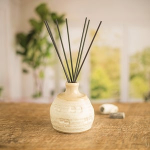 May include: A ceramic diffuser bottle with a white and beige glaze and a textured design. The bottle is filled with black diffuser sticks.