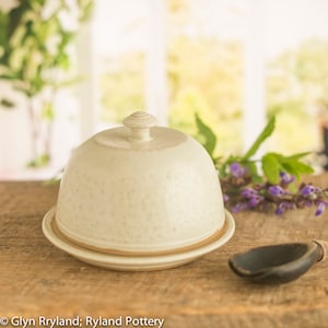 May include: A white ceramic butter dish with a lid and a small wooden spoon. The dish has a speckled glaze and a brown rim.