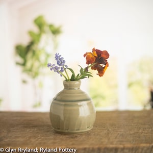 Może przedstawiać: Mały, jasnoszary ceramiczny wazon o zaokrąglonym korpusie i wąskiej szyjce, w którym znajduje się bukiet fioletowych i pomarańczowych kwiatów.