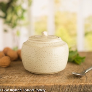 May include: A white ceramic jar with a lid, speckled with brown flecks. The jar is sitting on a wooden surface.