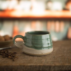 May include: A handcrafted ceramic mug, glazed in green on the upper half and natural, unglazed on the lower half. The mug features a rounded form and a robust handle. Coffee beans are scattered on the wooden surface near the mug.
