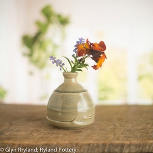 May include: A small, grey ceramic vase with a textured surface holds a bouquet of purple and orange flowers. The vase is sitting on a wooden table.