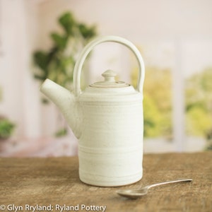May include: A white ceramic teapot with a rounded body and a lid. The teapot has a handle and a spout. A silver spoon is in the foreground.