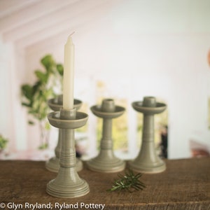 May include: Four grey ceramic candle holders with a single white candle in one of the holders. The candle holders are on a wooden surface.