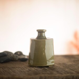 May include: A small, olive-green ceramic bottle with a square base and a rounded neck. The bottle is glazed and has a glossy finish. It sits on a wooden surface, with a blurred background.