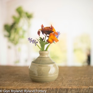 Könnte beinhalten: Eine kleine, hellgraue Keramikvase mit rundem Körper und schmalem Hals, die einen Strauß orangefarbener und lilafarbener Blumen hält.