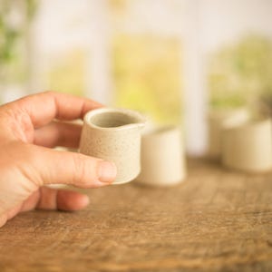 Op de afbeelding: Een kleine, witte keramische roomkan met een gespikkelde afwerking. De roomkan wordt in een hand gehouden.