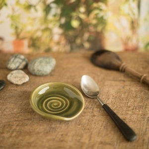 Könnte beinhalten: Ein kleiner, olivgrüner Keramik-Löffelhalter mit Spiralmuster, neben einem silbernen Löffel mit schwarzem Griff. Der Löffelhalter und der Löffel befinden sich auf einer Holzoberfläche, mit einem Holzlöffel und kleinen Steinen im Hintergrund.