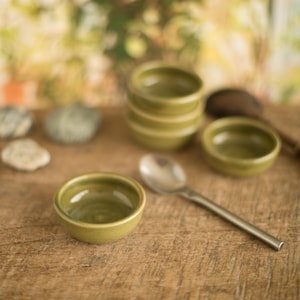 Könnte beinhalten: Eine Anordnung kleiner, olivgrüner Keramikschalen und ein silberner Löffel auf einer Holzoberfläche. Die Schalen sind gestapelt und verstreut, was auf eine Servier- oder Präsentationsanordnung hindeutet. Der Löffel befindet sich in der Nähe.