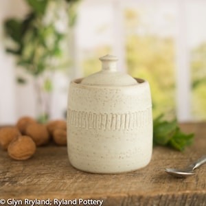 Könnte beinhalten: Weiße Keramik-Zuckerdose mit Deckel. Die Dose hat eine gesprenkelte Glasur und einen Band aus erhabenen Linien um die Mitte.