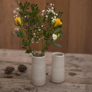 May include: Two white ceramic vases with a textured finish, one holding a bouquet of white and yellow flowers. The flowers include baby's breath, yellow roses, and white freesia.