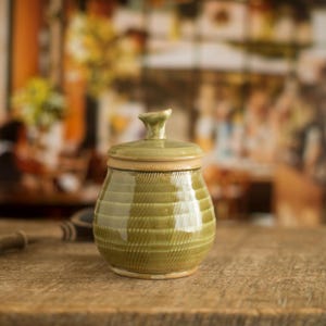 以下が含まれることがあります： 蓋付きの小さなオリーブグリーンのセラミックジャー。ジャーは、テクスチャード加工されたリブのデザインと、明るい茶色のリムが特徴です。蓋には小さなノブが付いています。ジャーは木製の表面に置かれています。