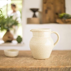 May include: A cream-colored ceramic pitcher with a handle and a spout, sitting on a wooden surface. The pitcher has a speckled texture and horizontal bands around the body. A small, matching bowl is in the foreground. The background is blurred.