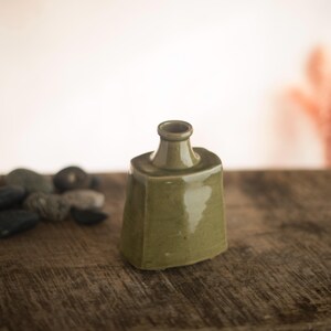 May include: A small, olive-green ceramic bottle with a geometric shape. The bottle has a square base and a cylindrical neck. It sits on a wooden surface, with a blurred background. The bottle is likely for decorative purposes.