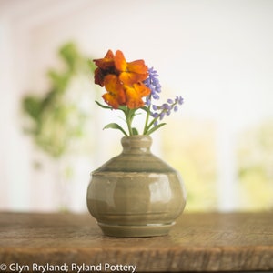 Peut inclure: Un petit vase en céramique gris avec un corps arrondi et un bord évasé contient un bouquet de fleurs orange et violettes.