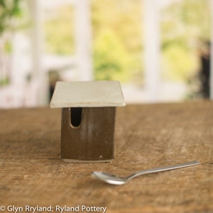 May include: A small, handcrafted ceramic house with a brown body and a cream-colored roof. The house has a rectangular shape with a cutout for a door. A silver spoon rests on the wooden surface next to the house. The background is blurred, suggesting a natural setting.
