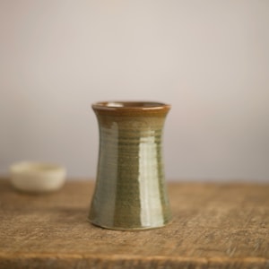 Può includere: Un vaso in ceramica fatto a mano con base svasata e collo stretto. Il vaso è smaltato in tonalità di verde e marrone, con una superficie strutturata. Il vaso è alto circa 15 cm ed è adatto per esporre fiori o come oggetto decorativo.