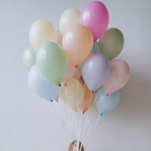 Peut inclure: Une jeune personne est assise sur une boîte en tenant un bouquet de ballons de couleur pastel. Les ballons sont attachés ensemble avec des rubans blancs.