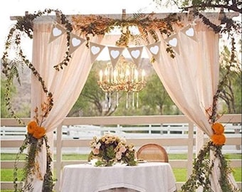 May include: A wedding ceremony setup with a white fabric canopy decorated with a burlap banner with white heart cutouts. The canopy is adorned with greenery and flowers. A white table with a white tablecloth is set up in front of the canopy.