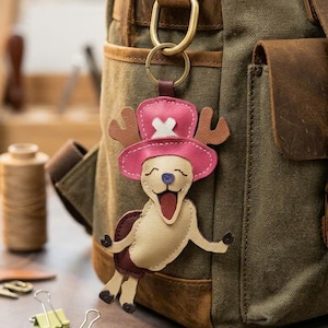 May include: A leather keychain shaped like a cartoon character wearing a pink hat with a white cross emblem, brown antlers, and a beige body with a purple nose and open mouth, attached to a brass-colored metal ring hanging from a green canvas backpack.