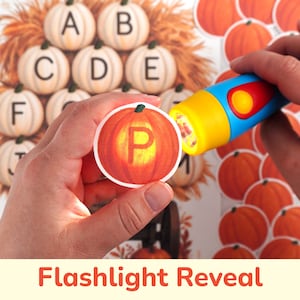 Op de afbeelding: Een ronde kaart met een oranje pompoen en de letter P in het midden, vastgehouden naast een blauwe en gele speelgoedzaklamp. Op de achtergrond is een vel papier te zien met meerdere pompoenen die zijn voorzien van letters.