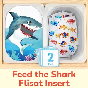 May include: A wooden toy with a shark image and a tray of colorful fish cards. The tray has a number card with the number "2" and the word "two" on it. The toy is called "Feed the Shark Flisat Insert".