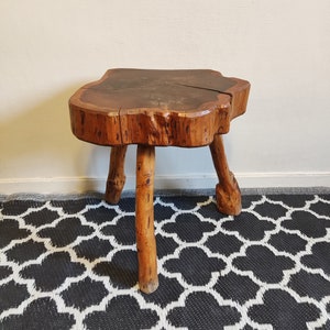 May include: A rustic wooden stool with a natural wood top and three legs. The stool is made from a tree trunk slice and has a rough, organic texture.