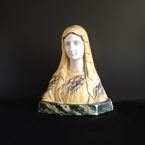 Puede incluir: Un busto de yeso blanco de la Virgen María con una túnica de patrones amarillos y marrones. El busto está sobre una base de mármol verde y negro.