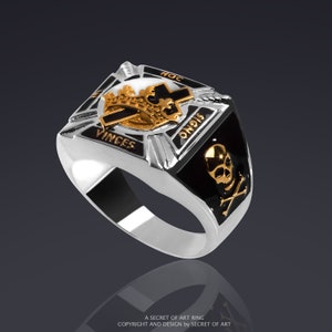 May include: A silver ring with a black enamel band. The ring features a gold crown, cross, and skull and crossbones design. The words "HOC", "VINCES", and "SIGNO" are inscribed on the ring.