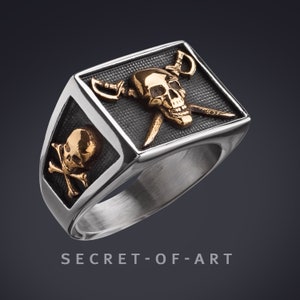 Op de afbeelding: Een zilveren ring met een zwart rechthoekig ontwerp met een gouden schedel met gekruiste zwaarden. De ring heeft ook een kleinere gouden schedel en gekruiste beenderen aan de zijkant. De tekst "SECRET-OF-ART" is onder de ring gedrukt.