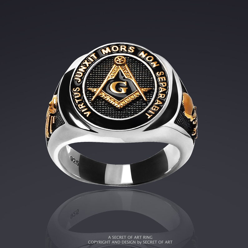 May include: A silver ring with a black enamel center featuring a gold Masonic compass and square with the letter 'G' inside. The ring has the inscription 'Virtus Junxit Mors Non Separabit' around the edge.