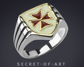May include: A silver ring with a gold shield-shaped top featuring a red cross with four points. The ring has a textured band.