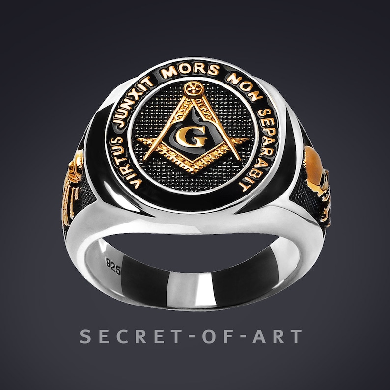 May include: A silver ring with a black enamel inlay featuring a gold Masonic compass and square with the letter "G" in the center. The ring is inscribed with the words "Virtus Junxit Mors Non Separabilit" in gold lettering.