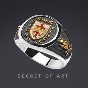 Könnte beinhalten: Ein silberner Ring mit einem schwarzen Emaille-Schädel und Knochen-Design. Der Ring hat außerdem ein vergoldetes Wappen mit einem roten Kreuz und einer Krone, umgeben von der Inschrift "IN HOC SIGNO VINCES".