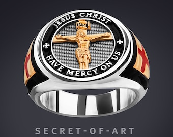 May include: A silver ring with a black and gold band. The center of the ring features a round, black enamel design with a gold cross and the words "Jesus Christ Have Mercy On Us".