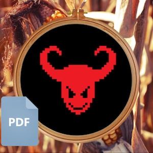 Puede incluir: Un cráneo de toro rojo pixelado con cuernos y una sonrisa amenazante sobre un fondo negro. El cráneo está en un aro de bordado de madera.