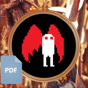 Puede incluir: Un diseño de arte de píxeles en blanco y negro de una criatura con alas rojas. La criatura es blanca con dos ojos negros y está de pie sobre un fondo negro. El diseño está en un aro de bordado circular.
