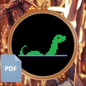 Puede incluir: Un patrón PDF en blanco y negro para bordar a punto de cruz una serpiente pixelada verde sobre un fondo negro. El patrón está en un aro de bordado redondo.