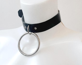 May include: A black leather choker necklace with a large silver ring pendant. The choker is secured with a buckle closure and is displayed on a white mannequin bust. The ring is approximately 2 inches in diameter.