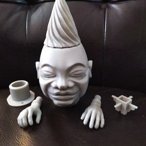 May include: A gray sculpture of a stylized human head with a cone-shaped hairstyle. The sculpture is in pieces, including a top hat, two hands, and a small square component. The pieces are all gray.