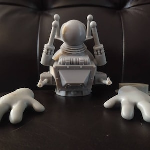 May include: A gray plastic robot toy with a round head, two arms, and a body with a rectangular chest piece. The robot has two antennas on top of its head and two arms that are separate from the body.