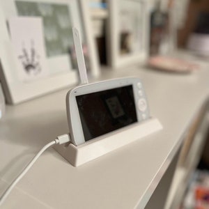 May include: A white baby monitor with a black screen and a white antenna. The monitor is sitting on a white stand and is plugged in with a white cord.