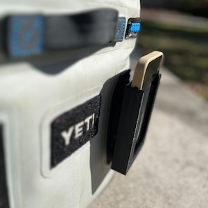 Puede incluir: Una nevera blanca Yeti con un soporte de tel&eacute;fono negro fijado al lateral. El soporte de tel&eacute;fono tiene una correa negra y una funda de tel&eacute;fono beige en el interior.