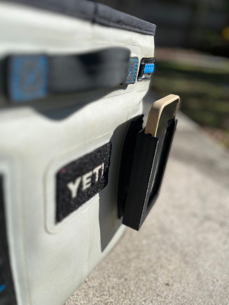 Puede incluir: Una nevera blanca Yeti con un soporte de tel&eacute;fono negro fijado al lateral. El soporte de tel&eacute;fono tiene una correa negra y una funda de tel&eacute;fono beige en el interior.
