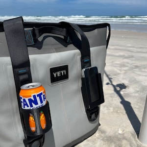 Puede incluir: Una bolsa de enfriador Yeti gris con una correa negra y un soporte para tel&eacute;fono negro. Una lata de Fanta Zero Sugar est&aacute; en un portavasos negro unido a la bolsa. La bolsa est&aacute; sentada en una playa de arena.