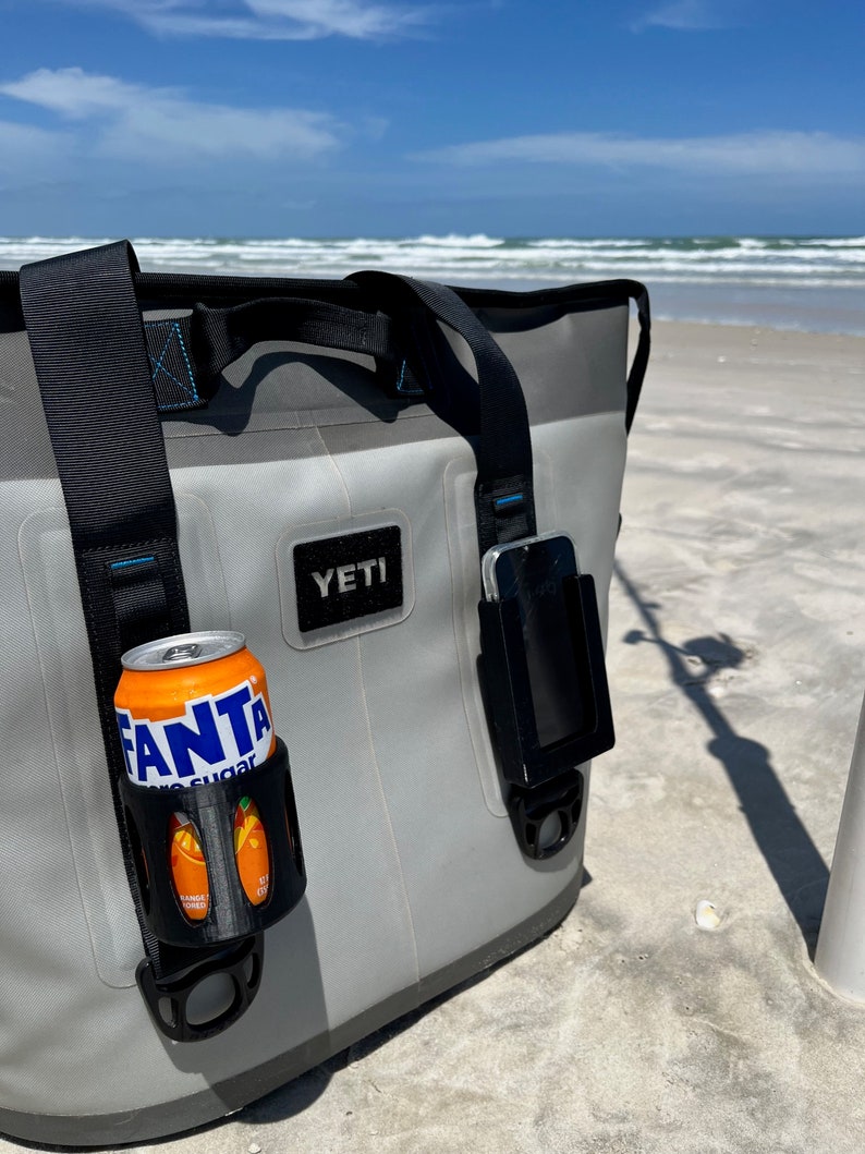 Puede incluir: Una bolsa de enfriador Yeti gris con una correa negra y un soporte para tel&eacute;fono negro. Una lata de Fanta Zero Sugar est&aacute; en un portavasos negro unido a la bolsa. La bolsa est&aacute; sentada en una playa de arena.