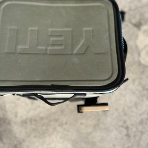 Puede incluir: Una bolsa de enfriador Yeti verde y negra con una correa negra y un asa de color beige. La bolsa tiene la palabra "YETI" en relieve en la parte delantera.
