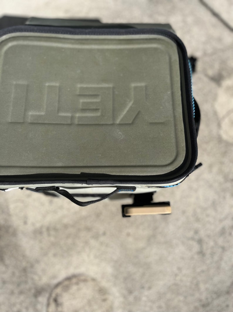 Puede incluir: Una bolsa de enfriador Yeti verde y negra con una correa negra y un asa de color beige. La bolsa tiene la palabra "YETI" en relieve en la parte delantera.