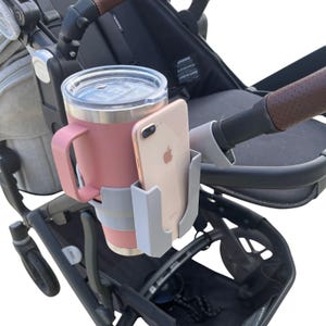 May include: A pink insulated tumbler with a handle and a silver lid is secured in a gray cup holder attached to a stroller. A smartphone is placed in a separate gray phone holder, also attached to the stroller. The stroller has a black frame and brown leather accents.